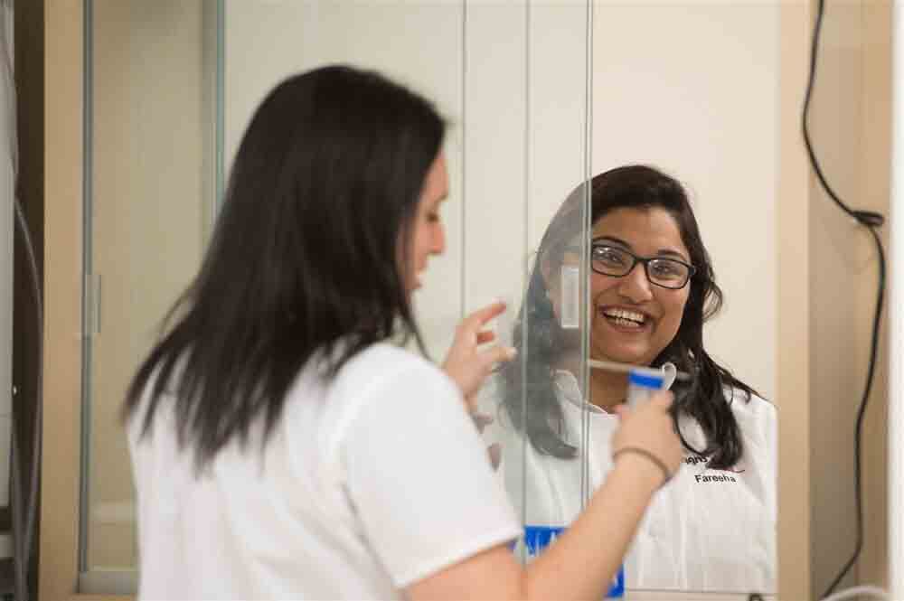 Two women in lab coats, one smiling in a mirror reflection, the other cleaning glass, creating a friendly, professional atmosphere.