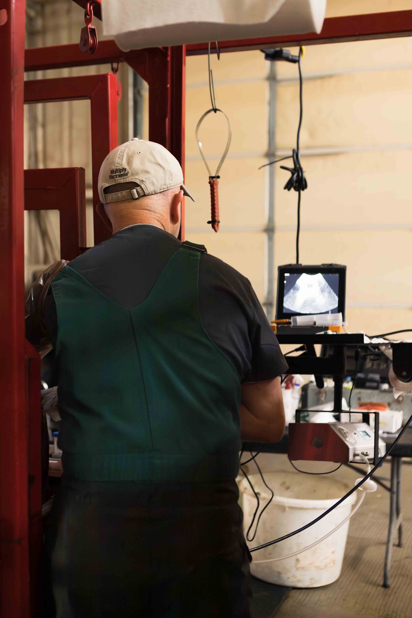 A man in a cap and apron examines an ultrasound monitor in a workshop, surrounded by equipment and wires.